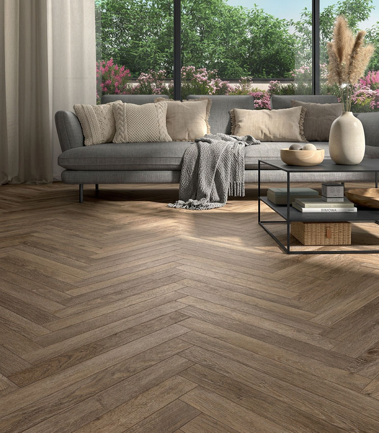 Contemporary living room with herringbone flooring from the Kairos Stigmi collection, furnished with a large grey sofa, metal coffee table, neutral-toned textiles and floor-to-ceiling windows overlooking a flowering garden.