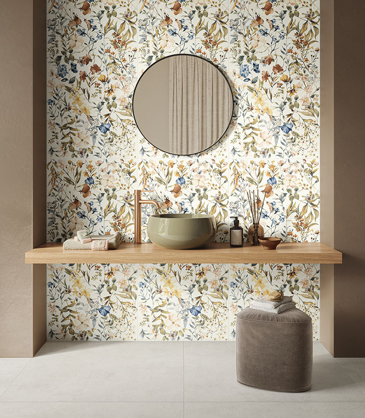 Contemporary bathroom featuring surfaces from the La Matière Talc and La Fleur collections, with a floral wall covering in natural tones, an olive green washbasin, wooden shelf, round mirror and upholstered pouf on light flooring.