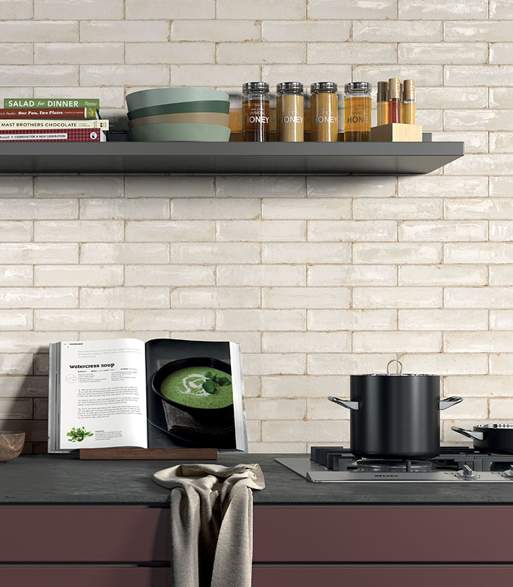 Detail of a contemporary kitchen featuring wall tiles from the Workshop Opal collection, with light rectangular tiles in an irregular glossy finish. The space includes dark shelves, a black worktop, burgundy cabinetry, cookbooks, containers and cookware.