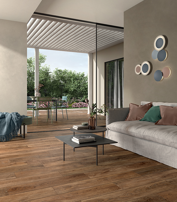Contemporary living room featuring flooring from the Kairos Zoi collection, with warm wood-effect surfaces, a large light-coloured sofa, low coffee tables and direct access to a furnished terrace overlooking the greenery.