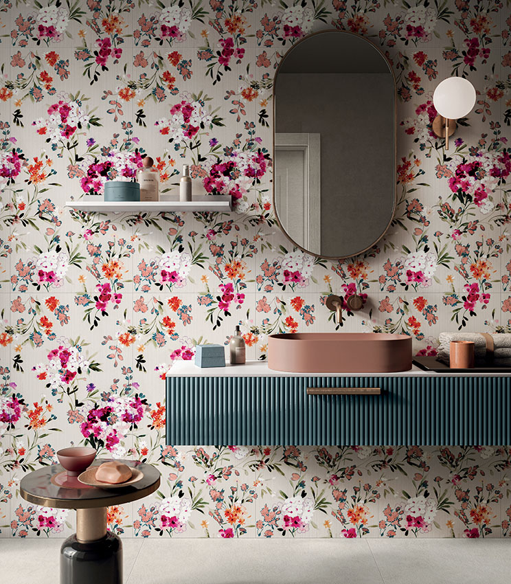 Contemporary bathroom featuring decorative wall covering from the Glam Bouquet collection, with a multicoloured floral pattern on a light background, a petrol blue floating vanity, dusty pink washbasin, large oval mirror and coordinated details in warm tones.
