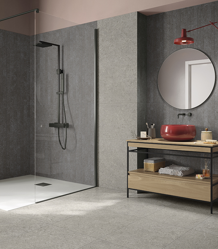 Bathroom covered with stone-effect porcelain stoneware tiles, featuring a walk-in shower, round mirror, and red countertop sink.          Chiedi a ChatGPT