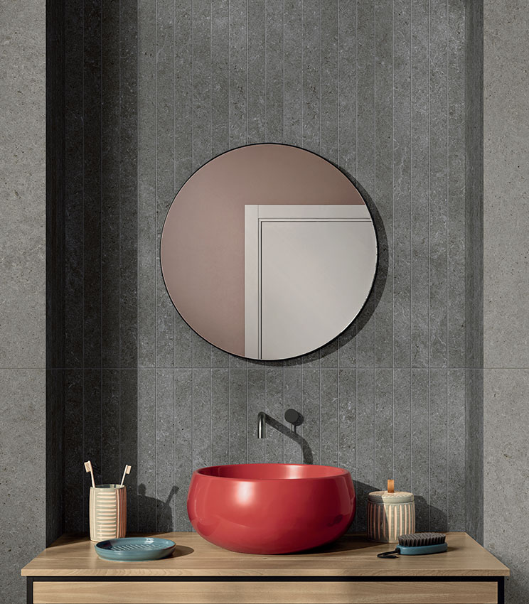 Bathroom covered with stone-effect porcelain stoneware tiles, featuring a walk-in shower, round mirror, and red countertop sink.          Chiedi a ChatGPT