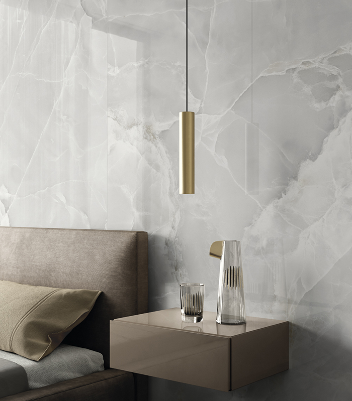 Detail of a wall covered with glossy, light marble-effect porcelain stoneware, featuring a floating bedside table, suspended lamp, and a glimpse of a double bed.