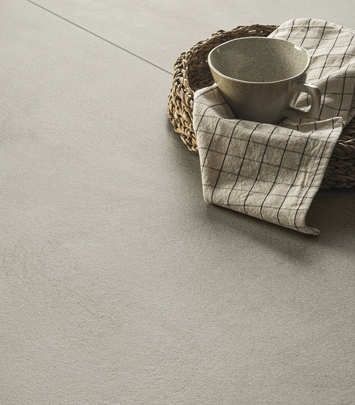 Scorcio ravvicinato di pavimento in gres effetto cemento di colore chiaro in grande formato, sul quale è posato un cesto di vimini contenente una tazza beige