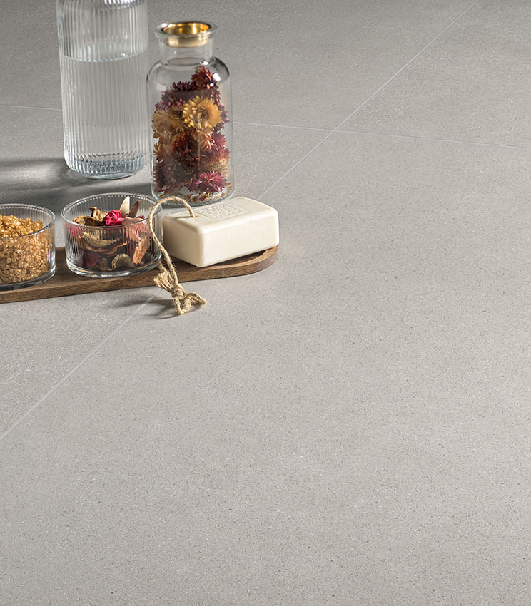 Detail of flooring from the Formula Form Ash collection, featuring a light, uniform surface with a textured appearance. The scene is completed by an arrangement with a glass bottle, a vase of dried flowers, small containers and a bar of soap on a wooden tray.