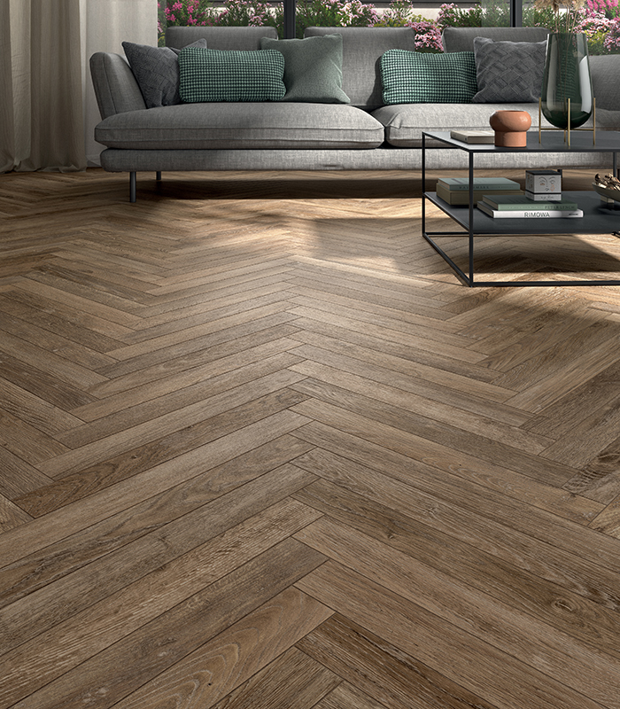 Scorcio di un living con un divano di tessuto grigio e una grande vetrata che affaccia su un giardino. Pavimento realizzato con listelli di gres porcellanato effetto legno posati a spina di pesce