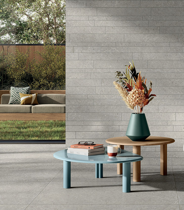 Contemporary living room featuring surfaces from the Surround Shape and Shape Muretto Soft collections, with grey stone-effect flooring and a wall clad in coordinated rectangular elements. The space is furnished with minimalist coffee tables, a vase with a floral arrangement and a wide opening onto a garden with an outdoor sofa.