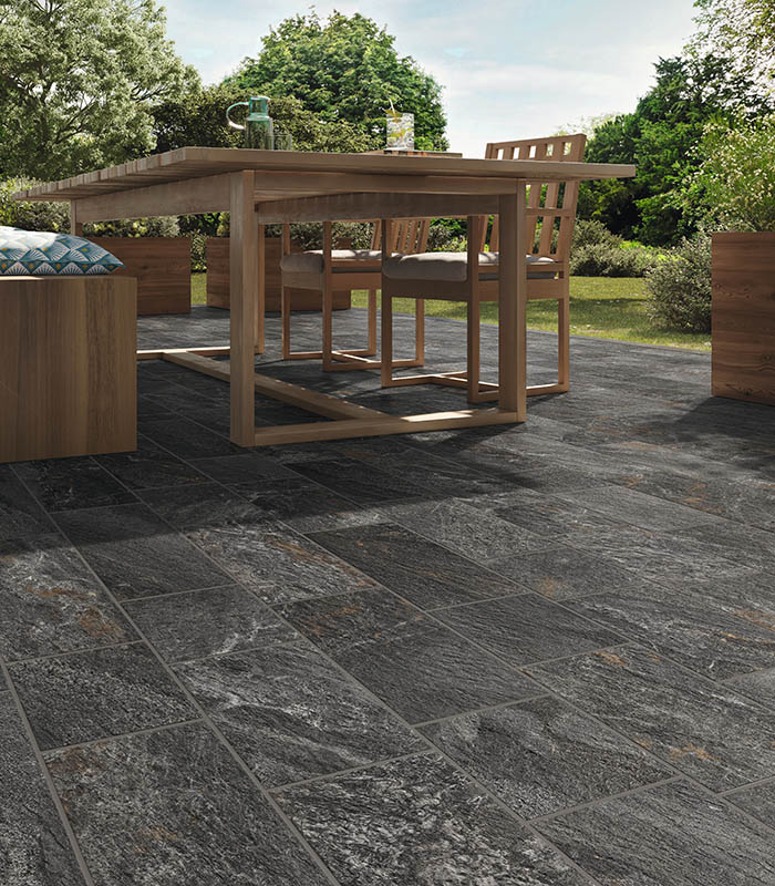 Vista ravvicinata di un pavimento outdoor effetto pietra di colore scuro posato con schema a correre multiformato. Tavolo e sedute in legno naturale di colore