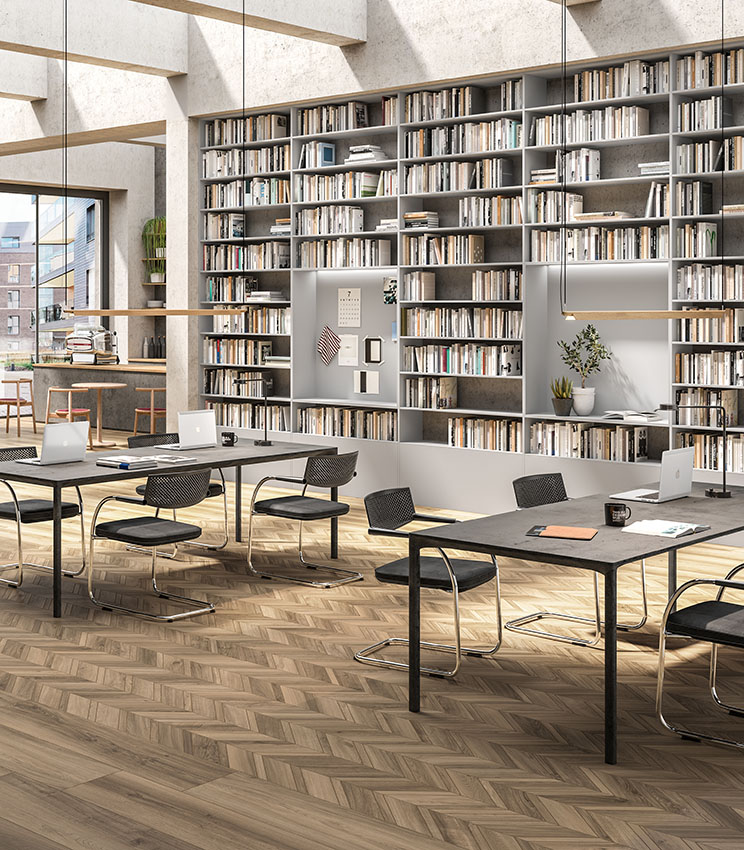 Contemporary library and workspace featuring flooring from the Nuance Cendre and Nuance Cendre Chevron collections, with wood-effect surfaces in natural tones and a geometric layout. The space includes large wall-to-wall bookcases, work tables with black chairs, laptops, decorative plants and large windows that fill the room with natural light.