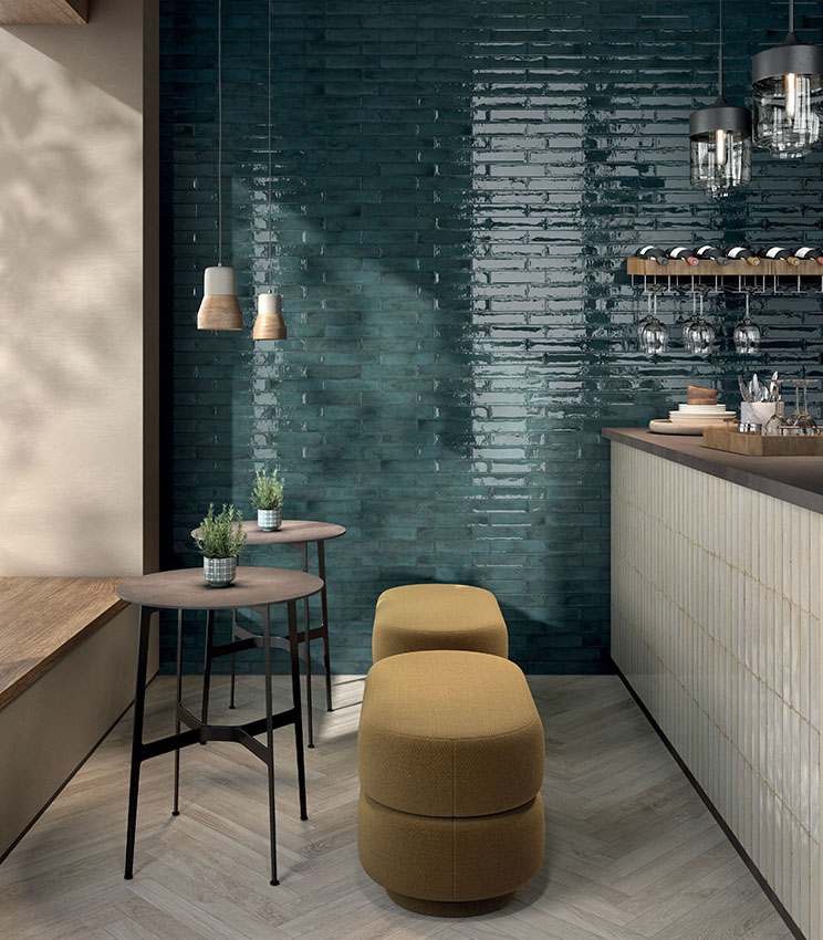 Contemporary bar corner featuring surfaces from the Workshop Berry, Workshop Opal and Kairos Imera collections, with a glossy petrol-green wall, a light-toned clad counter, wood-effect flooring and compact furnishings with upholstered stools and high tables.