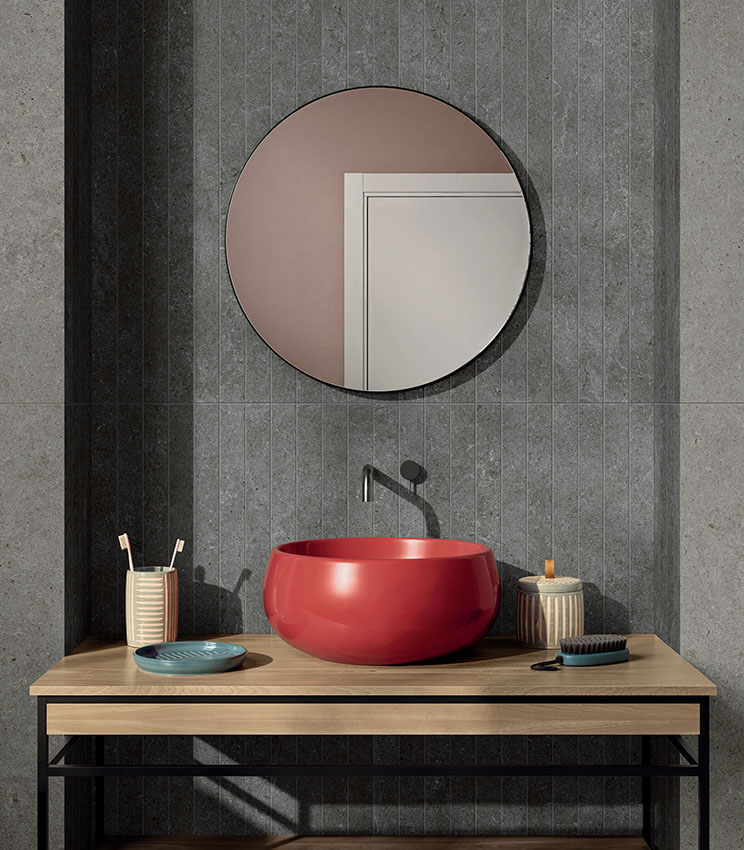 Contemporary bathroom featuring surfaces from the Surround Shape Soft and Outline Carving collections, with grey stone-effect coverings and a central section with a vertical linear pattern. The space features a glossy red washbasin on a wooden countertop, a round mirror, dark tapware and small bathroom accessories.