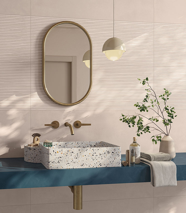 Contemporary bathroom featuring surfaces from the Lullaby Fairytale Blush and Linear Blush collections, with dusty pink wall coverings and bands with a linear relief texture. The space includes a rectangular terrazzo-effect washbasin, blue countertop, oval mirror with a gold frame, matching tapware, pendant light and decorative green branches.
