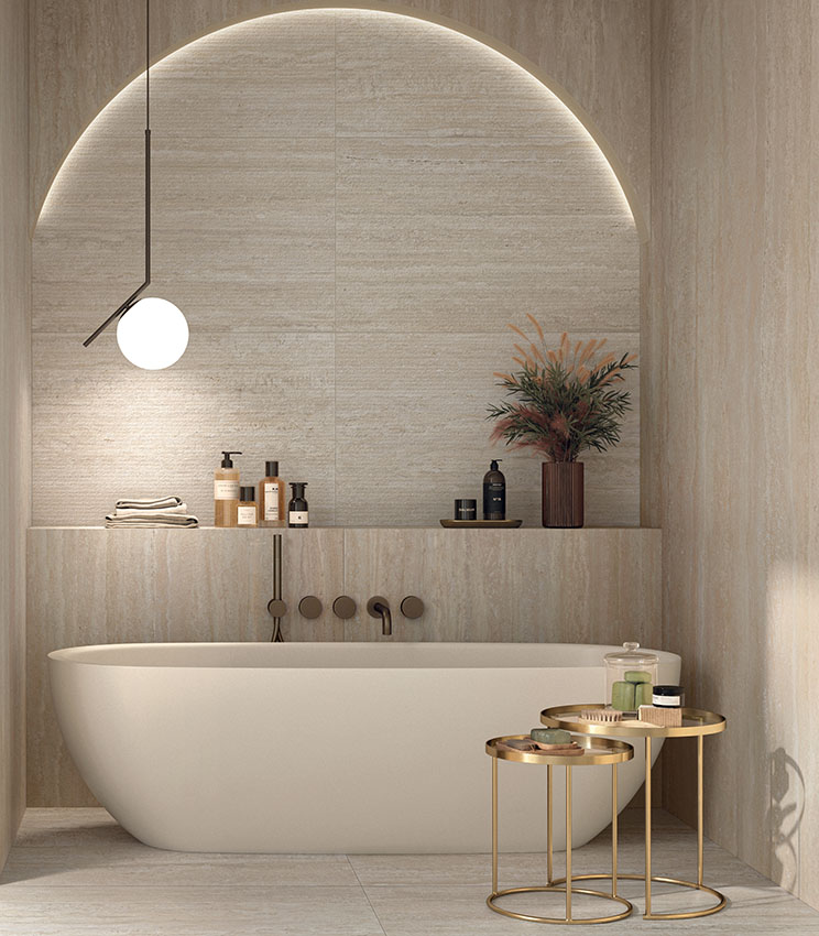 Contemporary bathroom featuring surfaces from the Perpetual Travertino Ivory Edge, Travertino Ivory and Travertino Cream collections, with travertine-effect finishes in warm, natural tones. The space includes a white freestanding bathtub set within a backlit arched niche, integrated shelves, a pendant light and gold-toned metal side tables with bathroom accessories.