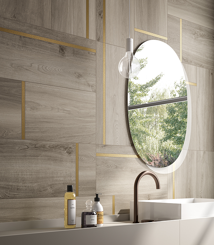 Detail of the washbasin wall in a small bathroom with ash-coloured wood-effect porcelain stoneware, featuring an oval mirror, a suspended light bulb, and tiles with a decorative brass insert.