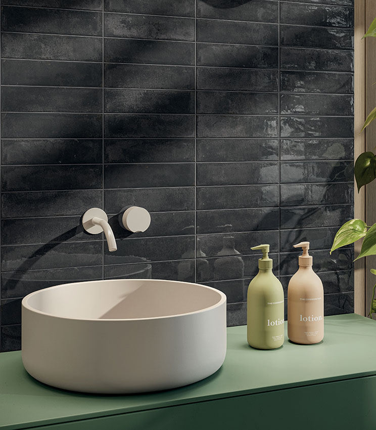 Detail of a contemporary bathroom featuring wall tiles from the Workshop Core collection, with dark rectangular tiles in a glossy, irregular finish. The scene includes a white countertop washbasin on a green vanity, wall-mounted tapware, coordinated bottles and a decorative plant.