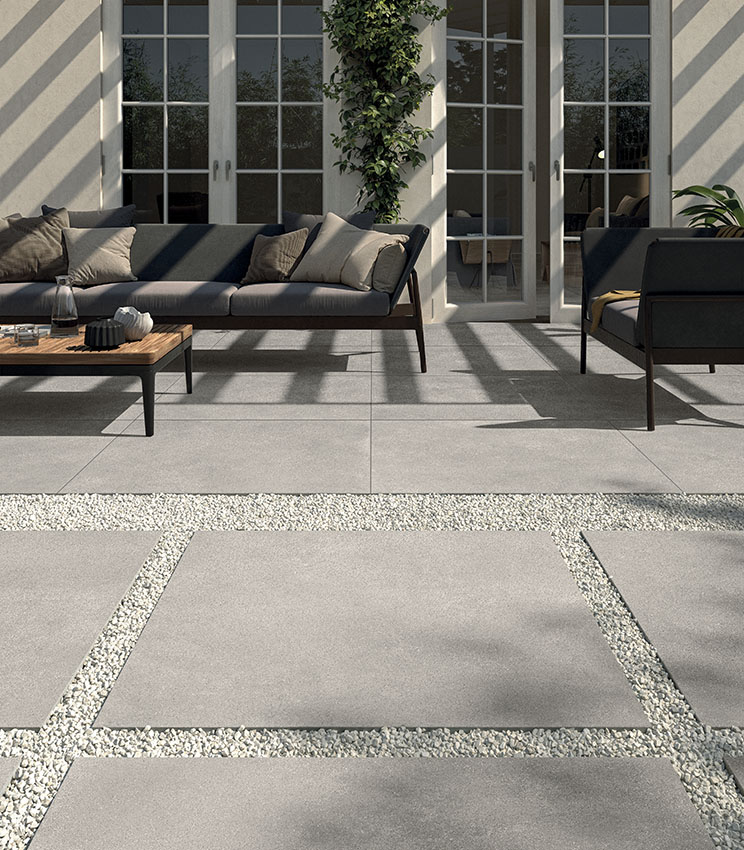 View of a patio furnished with a modern lounge area featuring a sofa and armchair. 20 mm concrete-effect porcelain stoneware flooring, installed with adhesive. In the foreground, an outdoor walkway made with large ceramic slabs dry-laid on gravel. | Formula collection by Panaria Ceramica