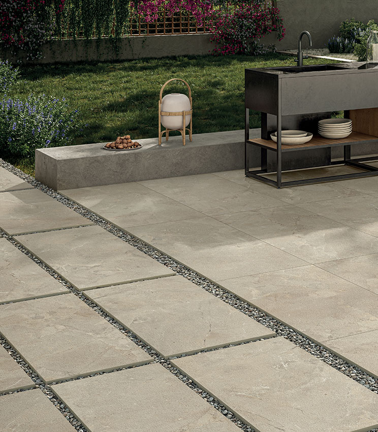 View of a poolside area with a modern outdoor kitchen unit and a green garden in the background. The porcelain stoneware flooring in sand-coloured stone effect is adhesive-laid around the pool and, in the foreground, dry-laid on gravel to form a pathway | Revel collection by Panaria Ceramica