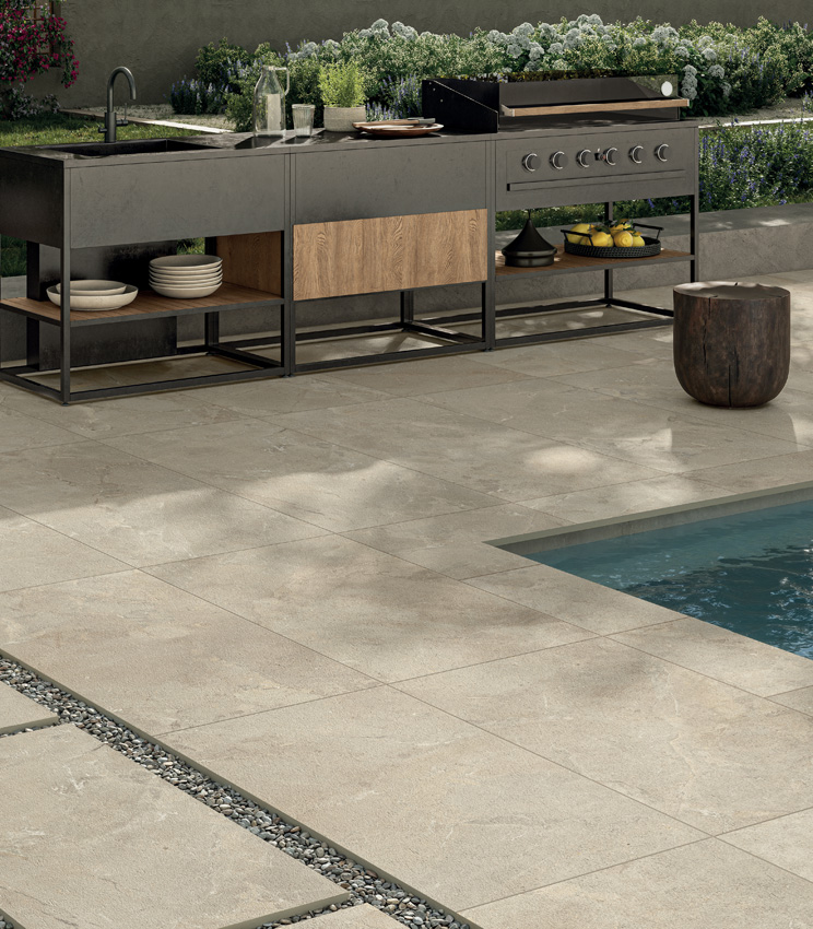 View of a poolside area with a modern outdoor kitchen unit and a green garden in the background. The porcelain stoneware flooring in sand-coloured stone effect is adhesive-laid around the pool and, in the foreground, dry-laid on gravel to form a pathway | Revel collection by Panaria Ceramica