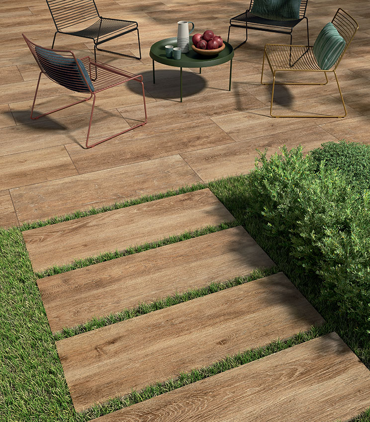 View of a patio with modern wire-frame chairs and a 20 mm wood-effect porcelain stoneware floor laid with adhesive. In the foreground, an outdoor ceramic plank pathway begins, dry-laid on grass | Kairos collection by Panaria Ceramica