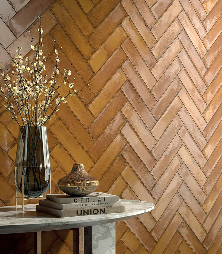 Detail of a wall clad with the Workshop Spice collection, featuring rectangular tiles laid in a herringbone pattern in warm terracotta tones. In the foreground, a marble table with books, decorative vases and flowering branches completes the setting.