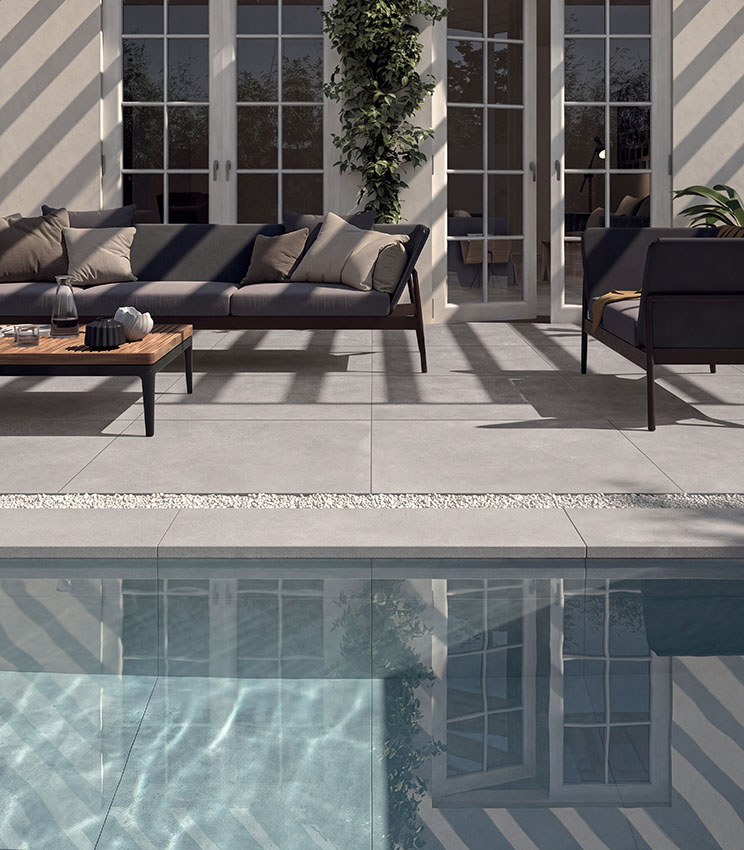 Poolside area with concrete-effect porcelain stoneware flooring, contemporary outdoor furniture and large windows, creating an elegant and functional space.